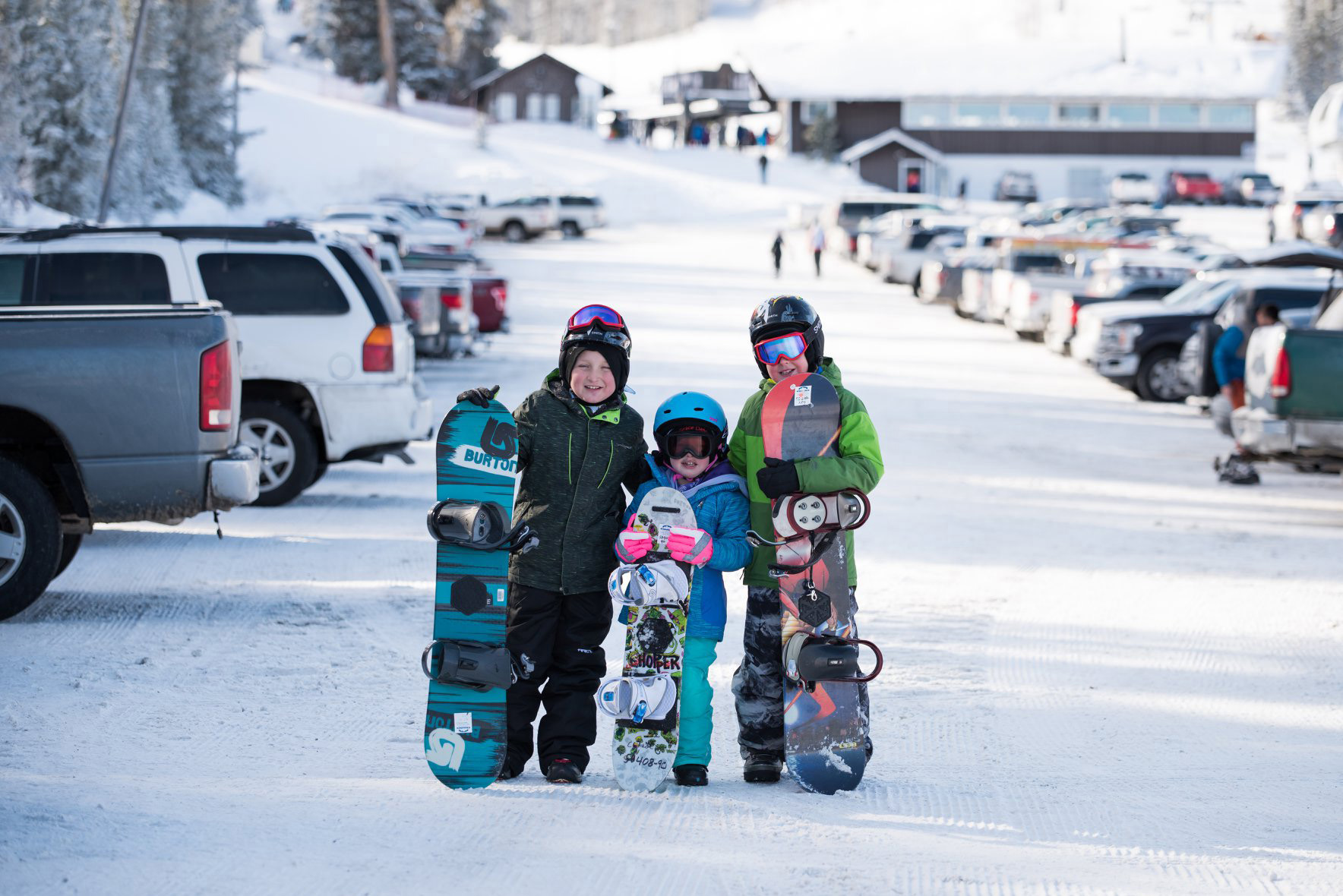 3 children holding snowboards pose in the snowy parking lot of Pomerelle Ski Area