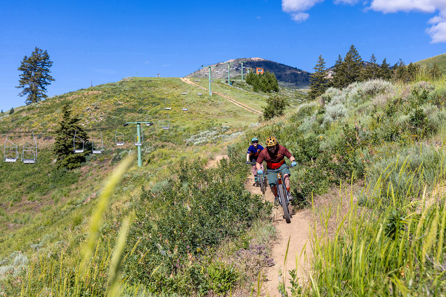 Two mountain bikers ride a dirt trail at Soldier Mountain in Idaho, passing beneath a ski lift on a grassy hillside with forested peaks in the background.